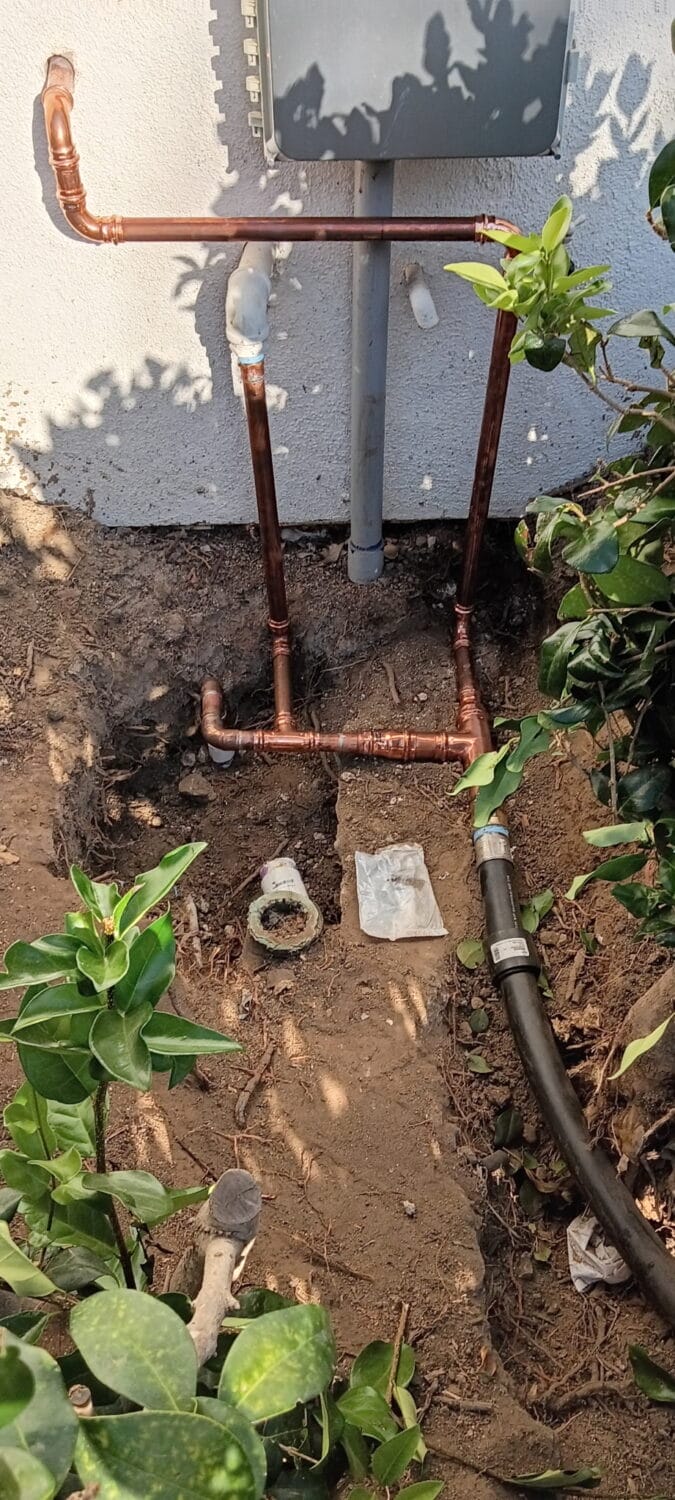 Copper and plastic pipes are installed vertically and horizontally along an outdoor wall, surrounded by soil, plants, and exposed plumbing fittings—a clear indication of recent repiping work to upgrade the plumbing system.