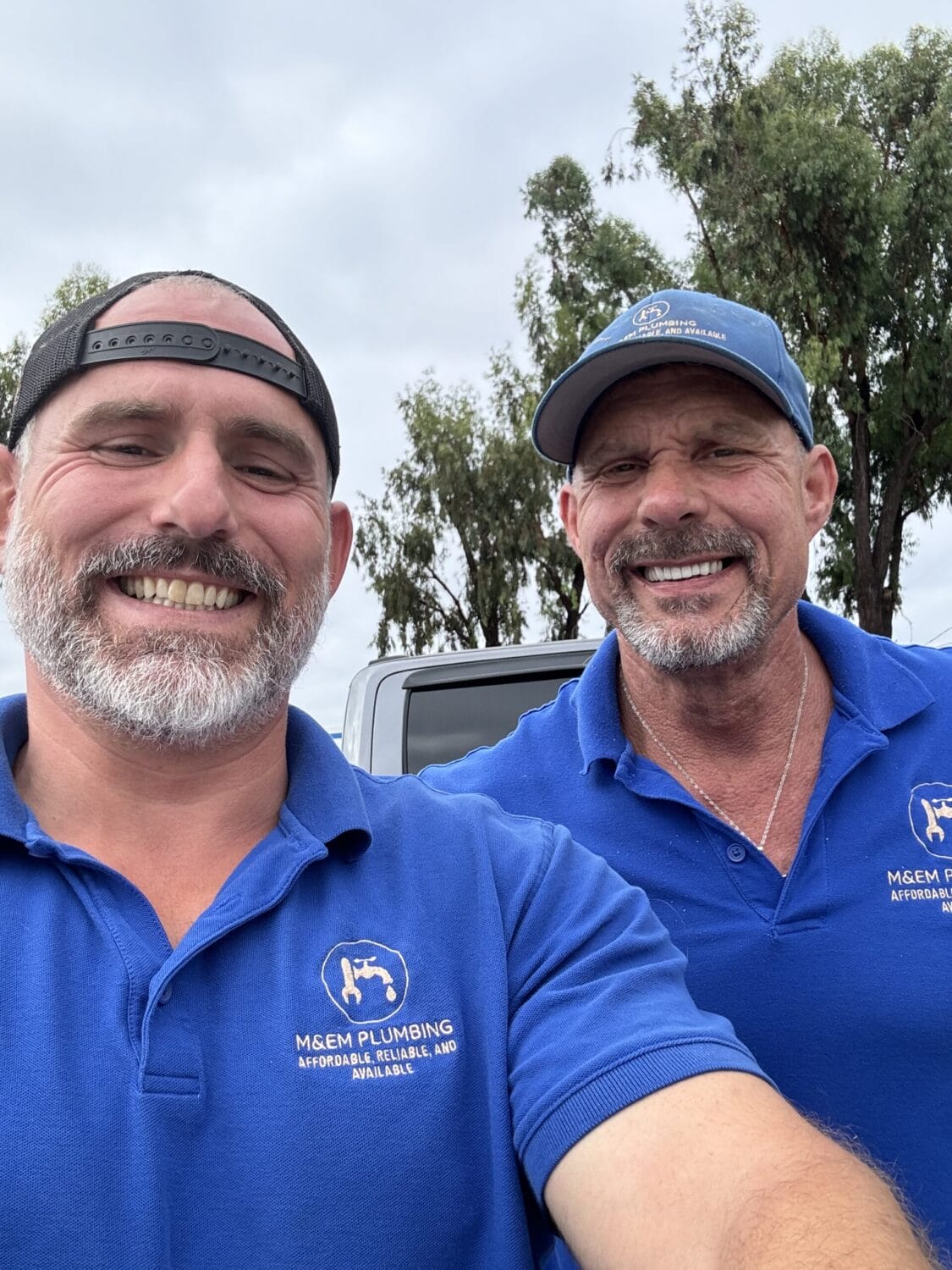 Two men in blue MSem Plumbing uniforms smile outdoors in front of trees and a vehicle.