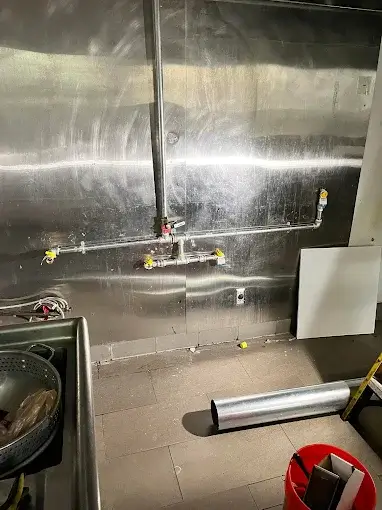 Commercial kitchen space with exposed metal pipes and fittings against a stainless steel wall, some construction materials and tools visible on the floor.