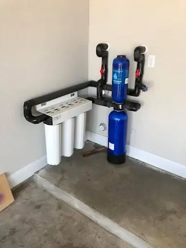 A water filtration system with three white filter housings and a blue tank is installed in the corner of a room with black piping attached to the wall.