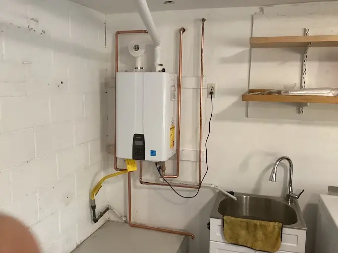 A tankless water heater is mounted on a white basement wall with visible copper pipes, next to a utility sink and wooden shelves.