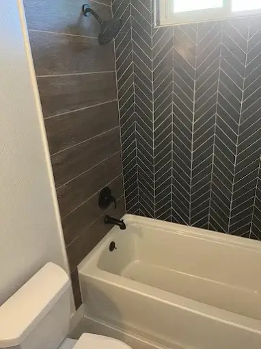 Modern bathroom with a white bathtub, black fixtures, and contrasting tile walls—one with wood-look tiles and the other with black herringbone tiles under a window.
