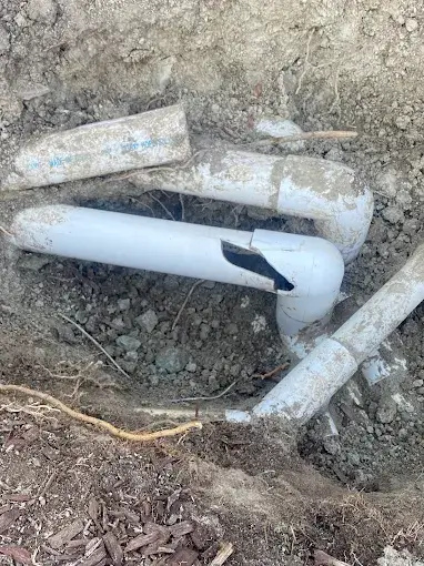 Broken white PVC pipes are partially buried in dirt, with one pipe clearly cracked open, exposing the inside.