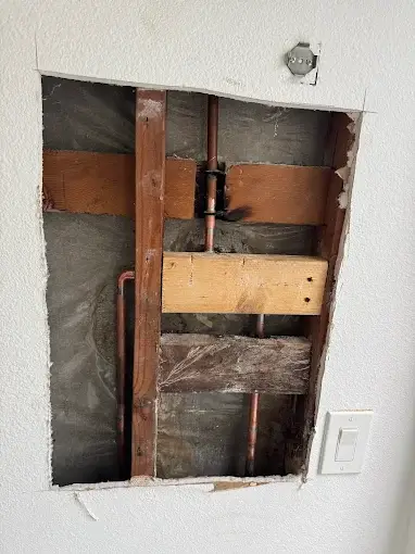 A section of drywall has been removed from a wall, exposing wooden studs, copper pipes, and an electrical outlet at the bottom right.
