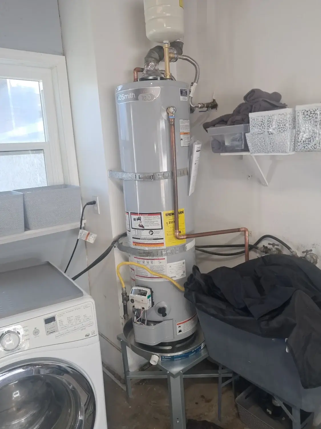 A water heater with attached pipes stands in a laundry room next to a washing machine, shelves, and a plastic bin, showcasing a seamless water heater installation that keeps the space organized and functional.